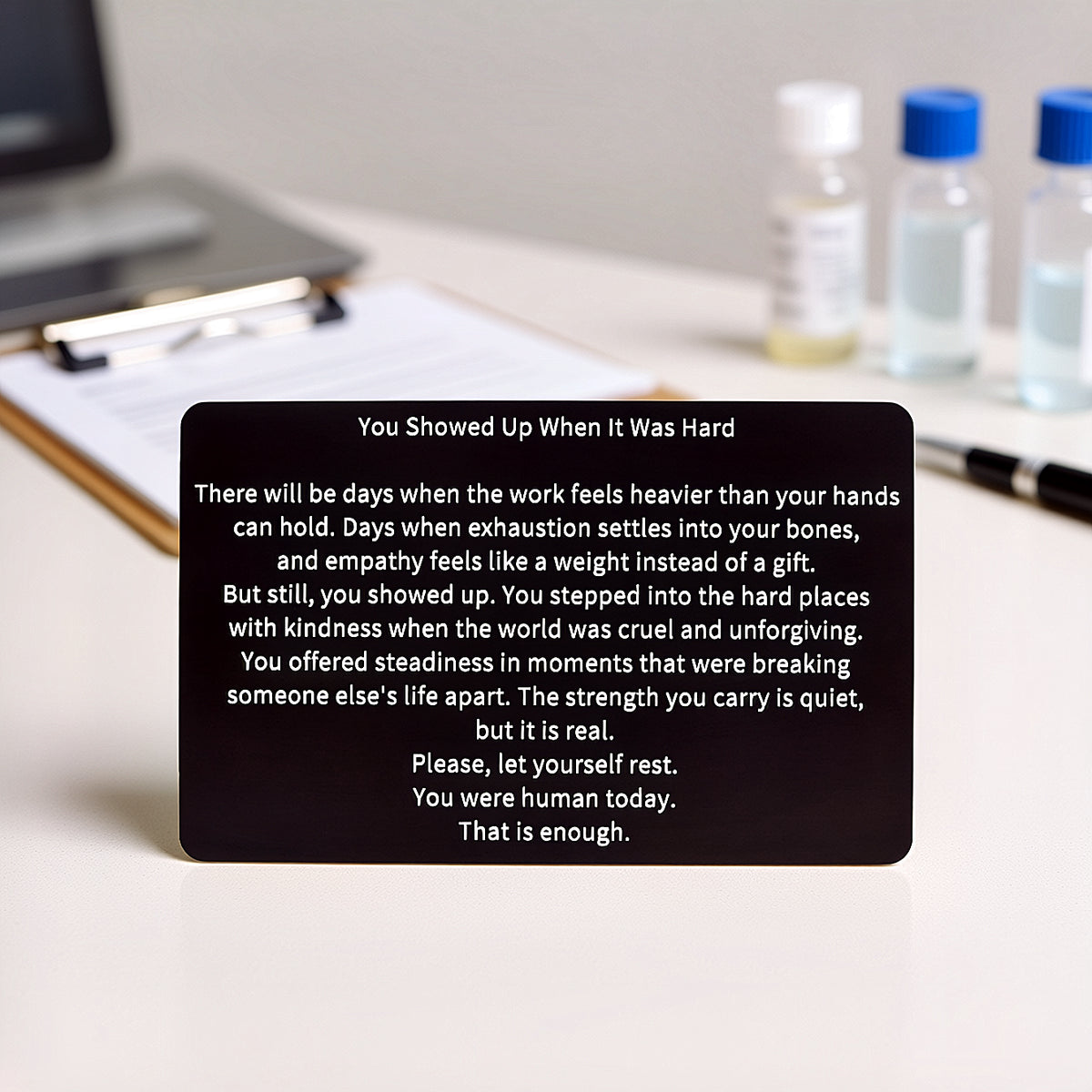 Black engraved metal wallet card reading 'You Showed Up When It Was Hard' lying on a white table with a clipboard with paper, a black pen, and three medical vials in the background.