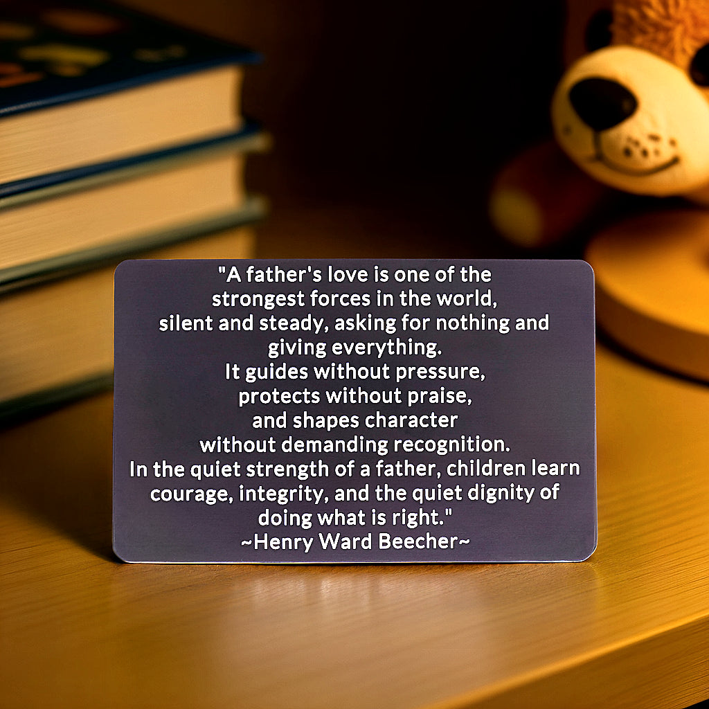 Black engraved metal wallet card with Henry Ward Beecher's quote on a father's love, standing on a wooden table with books and a teddy bear behind it.