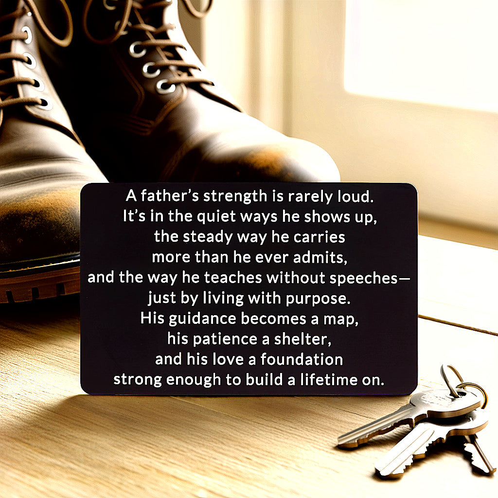 Black engraved metal wallet card with a message about a father's quiet strength, lying on a wooden floor next to worn leather work boots and a set of keys.