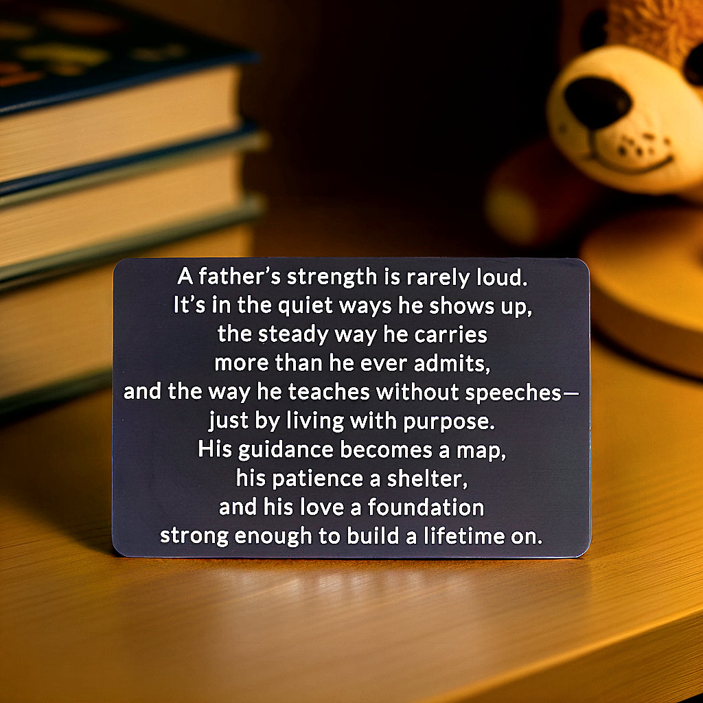 Black engraved metal wallet card with a message about a father's quiet strength, standing on a wooden table with books and a teddy bear behind it.