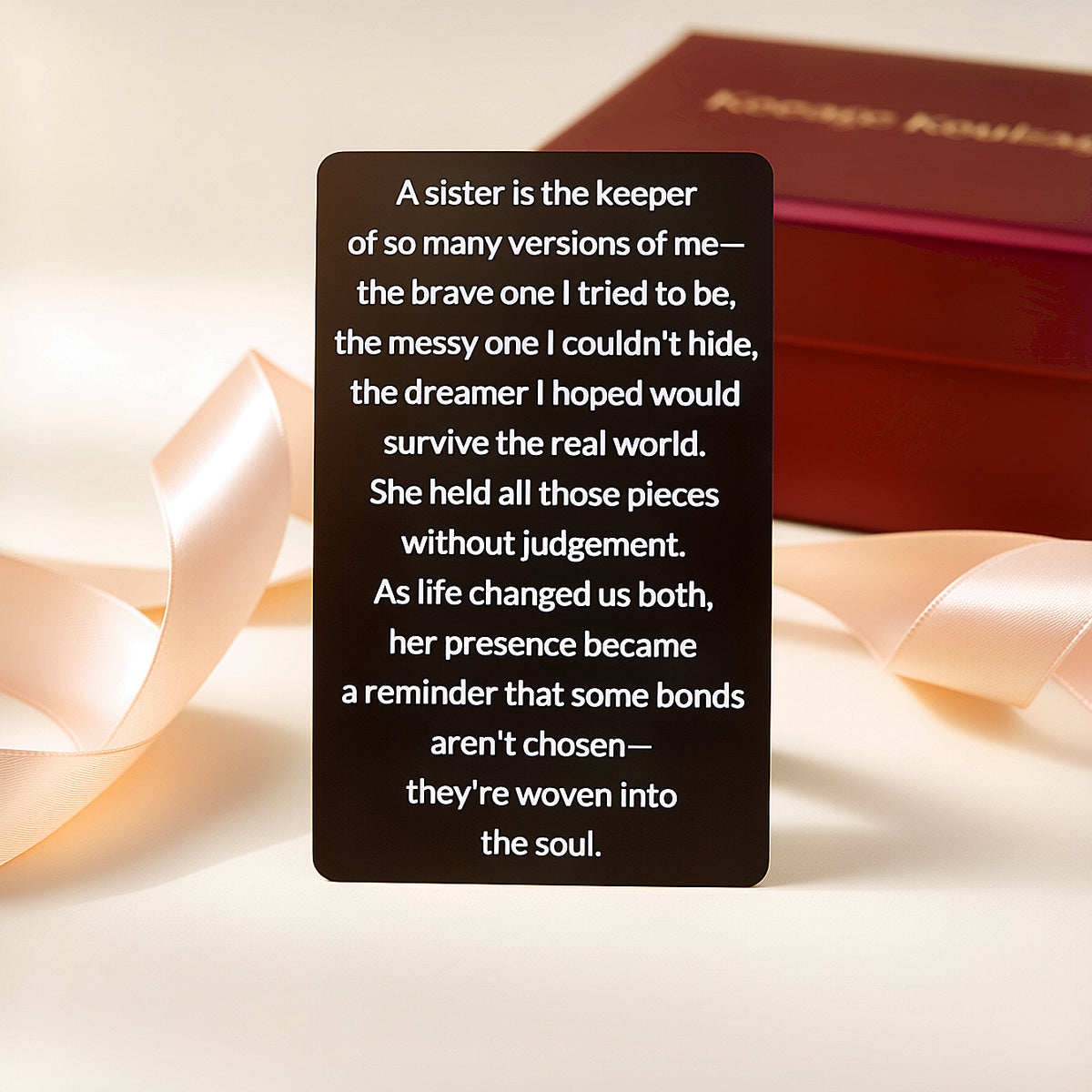 Black engraved metal wallet card with a message about a sister who holds every version of you, standing next to a red gift box and peach satin ribbon.