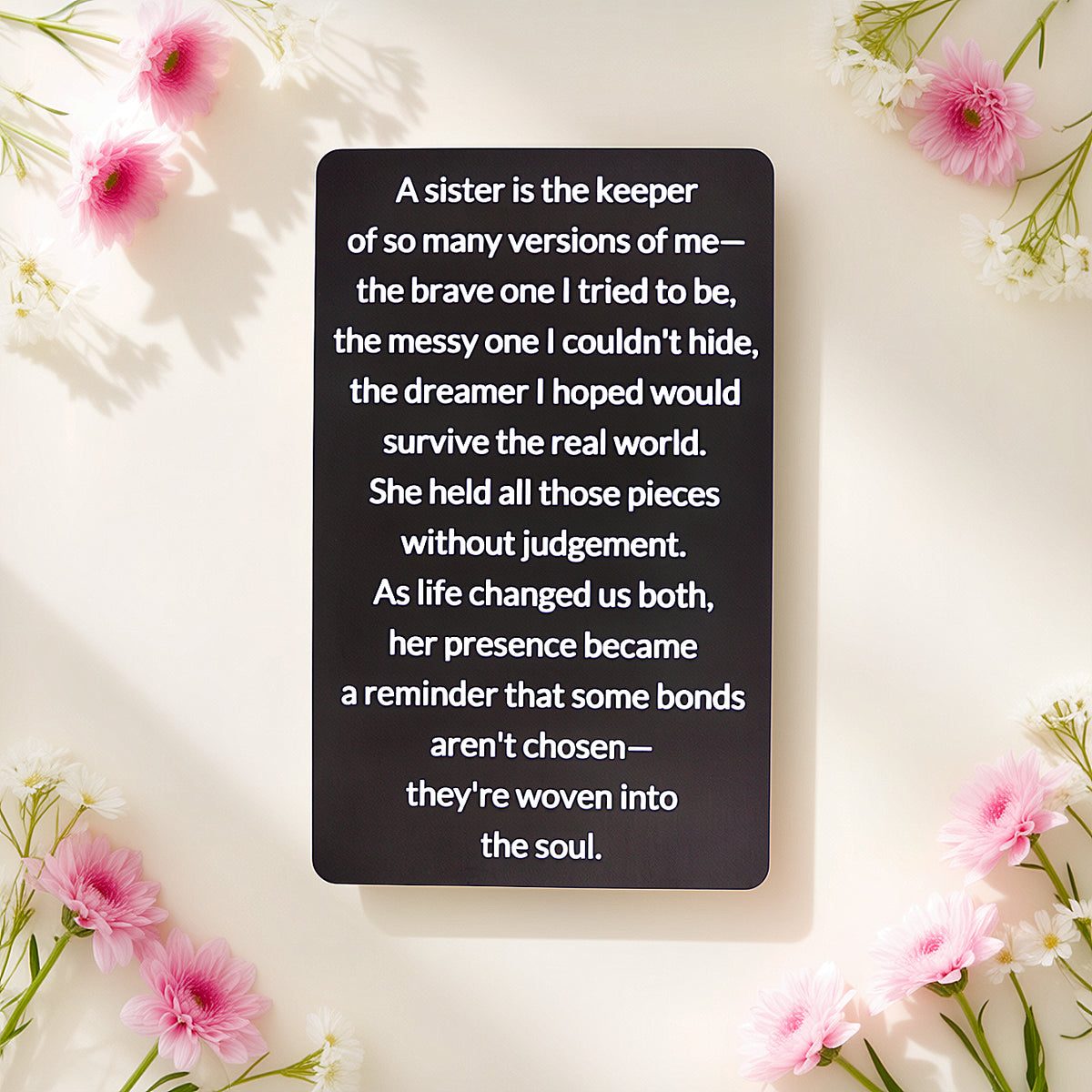 Black engraved metal wallet card with a message about a sister who holds every version of you, lying on a cream background framed by pink flowers.