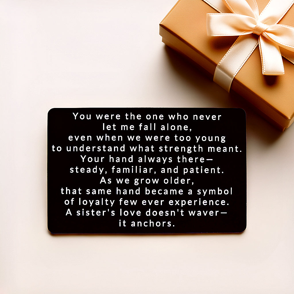 Black engraved metal wallet card with a message about a sister's love that anchors you, lying on a cream background next to a gold gift box with a satin bow.