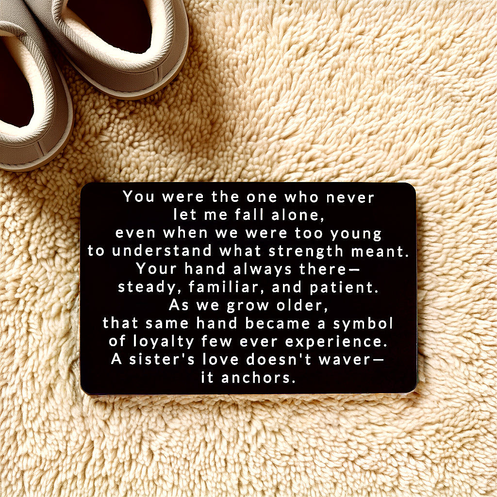 Black engraved metal wallet card with a message about a sister's love that anchors you, lying on a cream shag rug next to a pair of slippers.