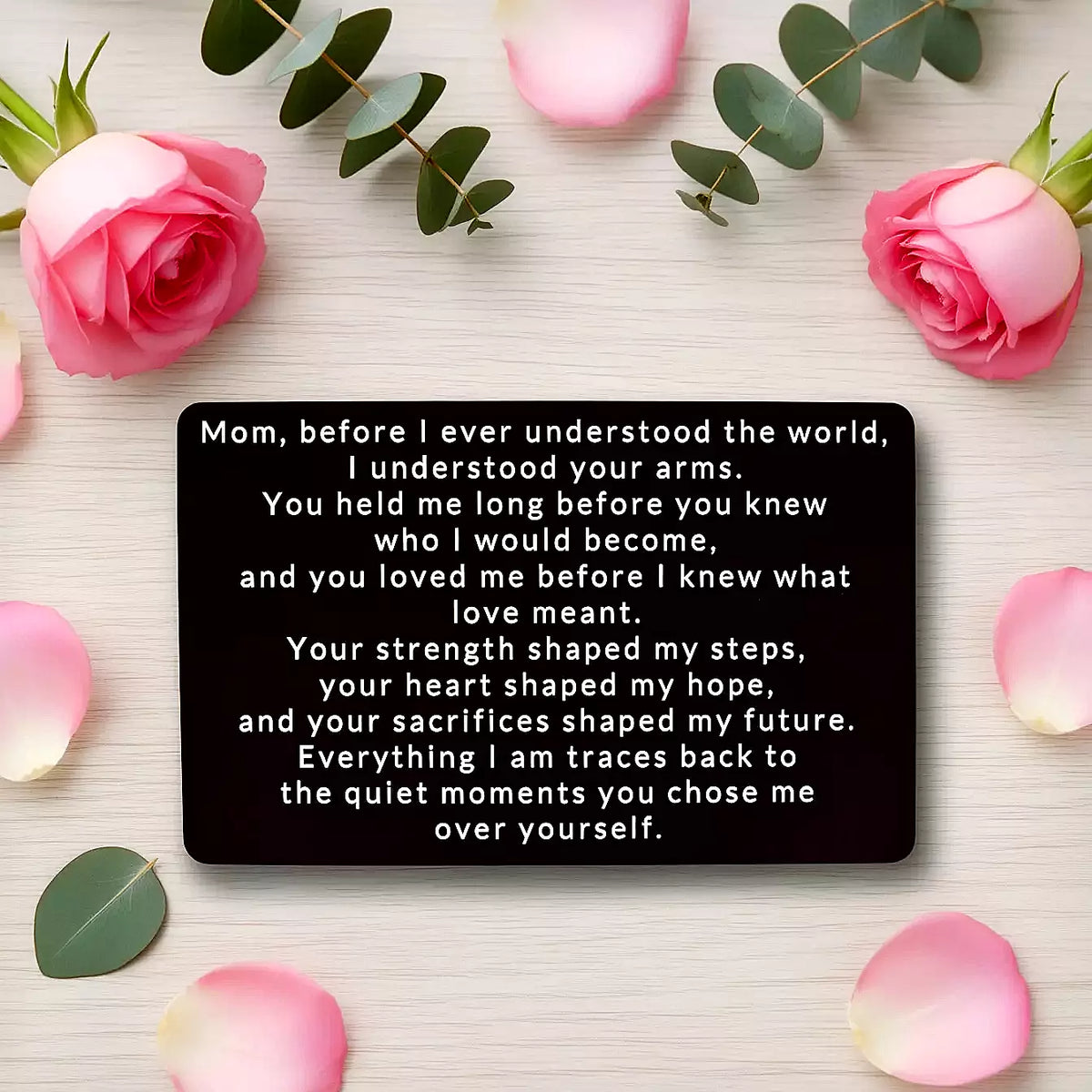 Black engraved metal wallet card reading 'Before I Ever Understood the World' on a wooden table surrounded by pink roses and rose petals.