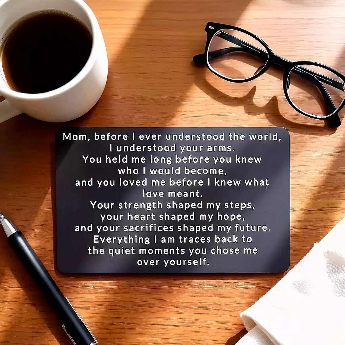 Black engraved metal wallet card reading 'Before I Ever Understood the World' on a wooden table next to a cup of coffee, glasses, and a pen.