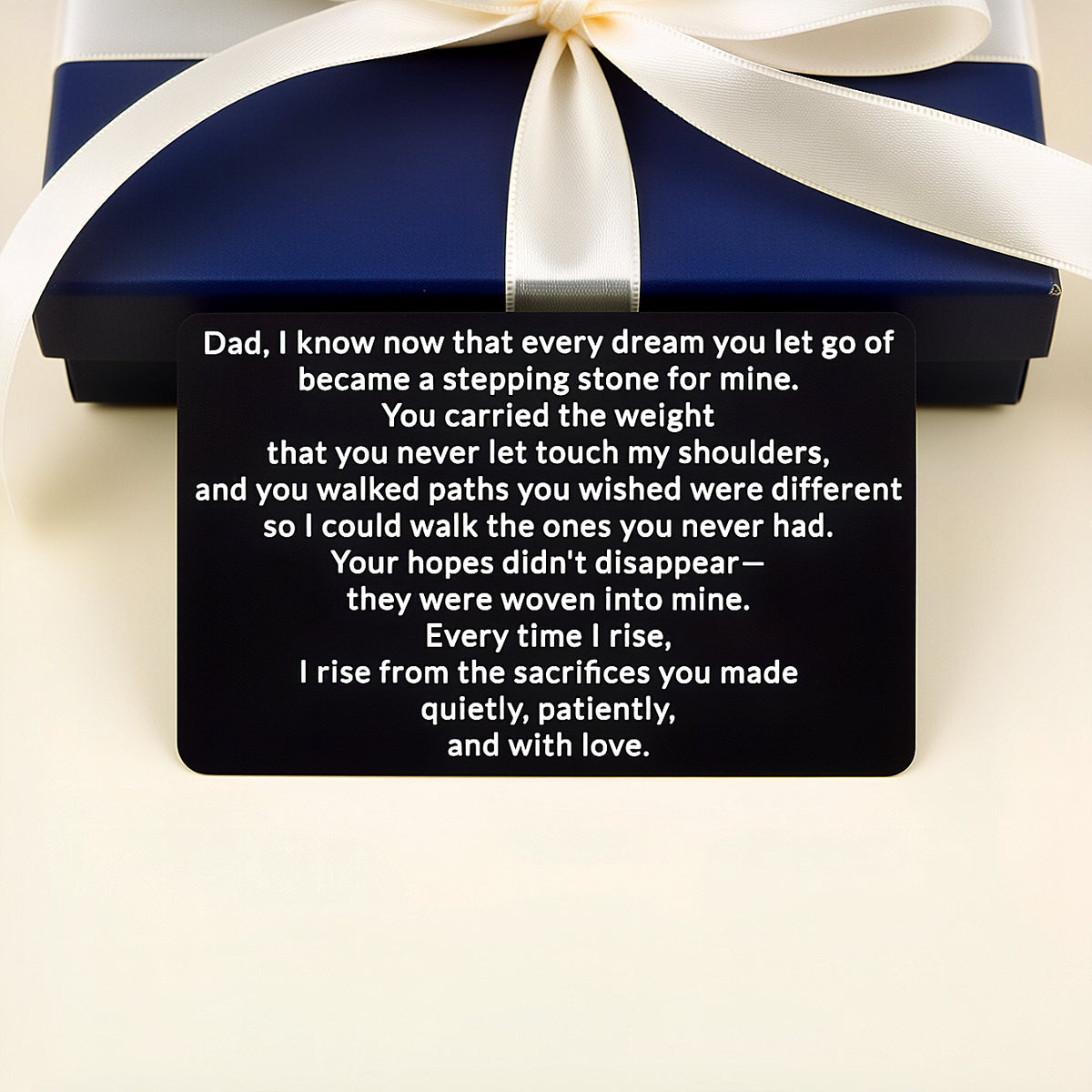 Black engraved metal wallet card with a message to Dad about the dreams he let go becoming stepping stones, lying in front of a navy gift box with a white ribbon.
