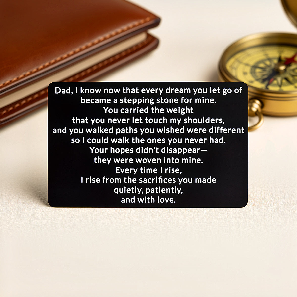 Black engraved metal wallet card with a message to Dad about the dreams he let go becoming stepping stones, standing in front of a brown leather wallet and a brass compass.