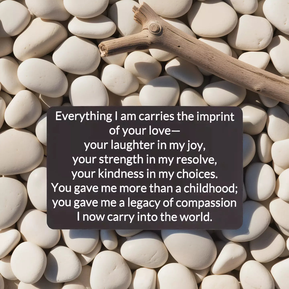 Black engraved metal wallet card reading 'Everything I Am Carries the Imprint of Your Love' lying on white river rocks and a wooden stick.