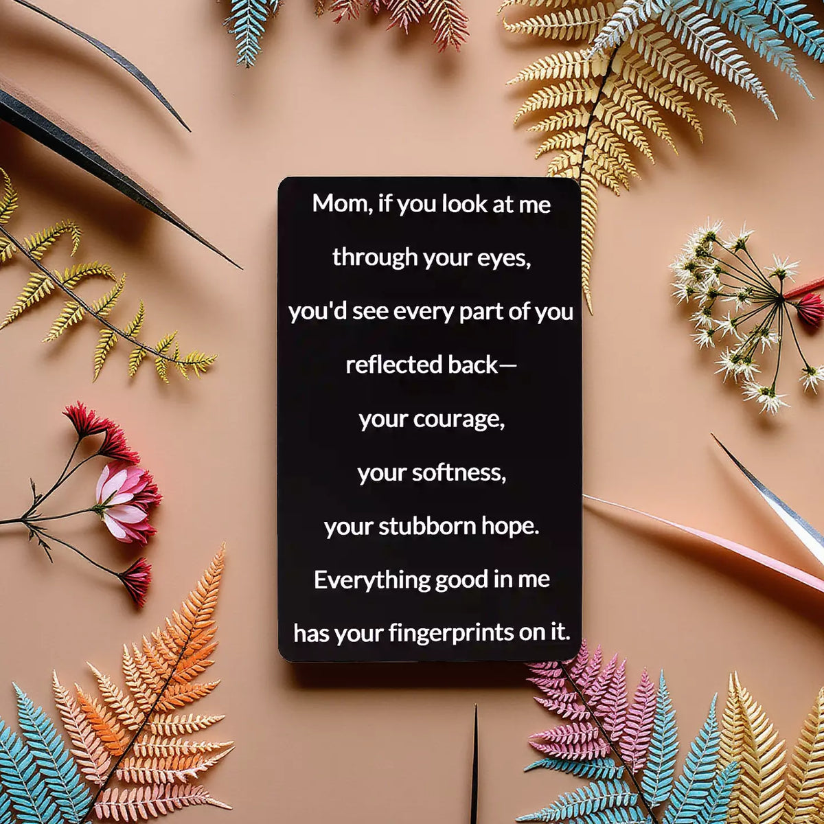 Black engraved metal wallet card reading 'Everything Good In Me Has Your Fingerprints' lying on a brown table surrounded by wildflowers
