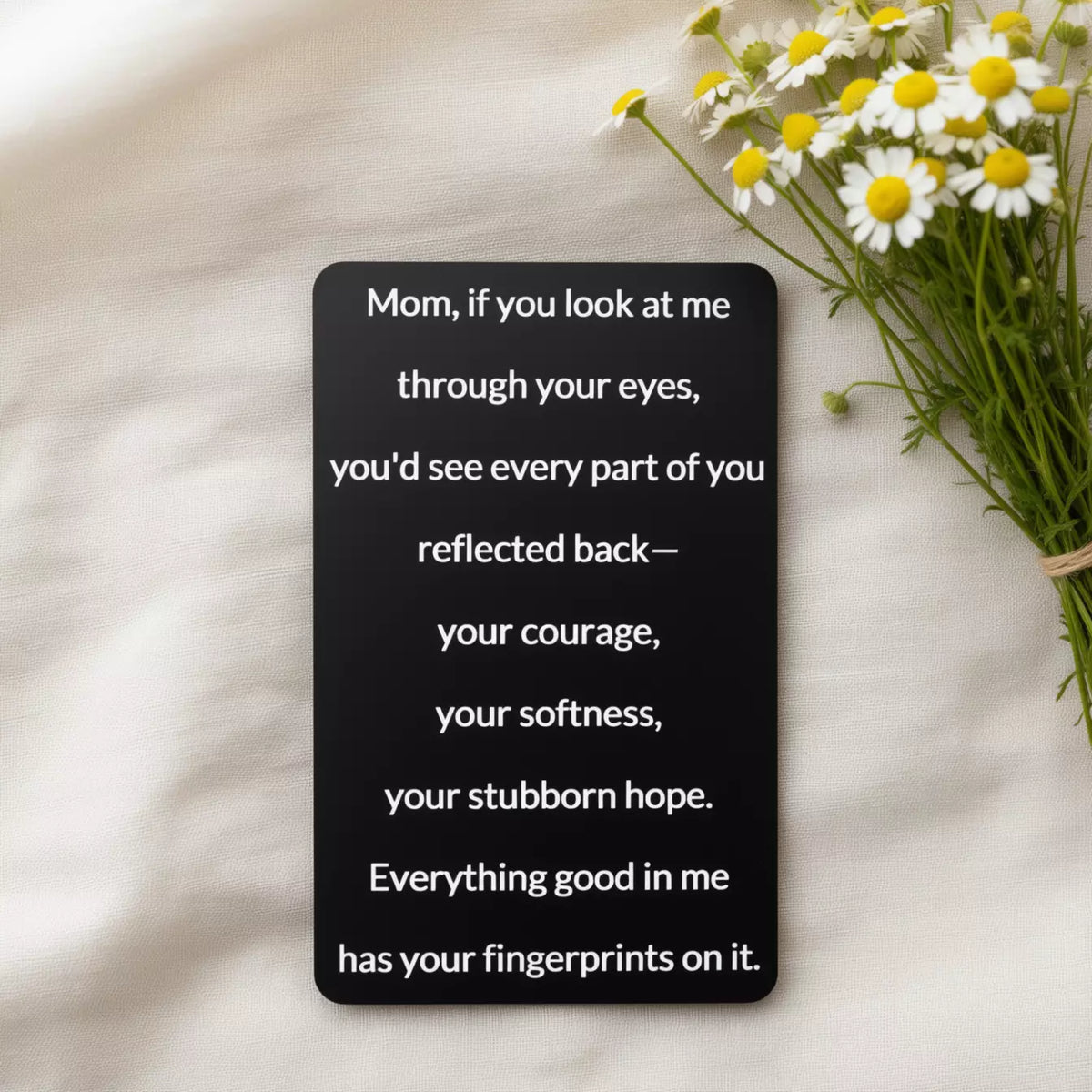 Black engraved metal wallet card reading 'Everything Good In Me Has Your Fingerprints' lying on white linen next to wildflowers.