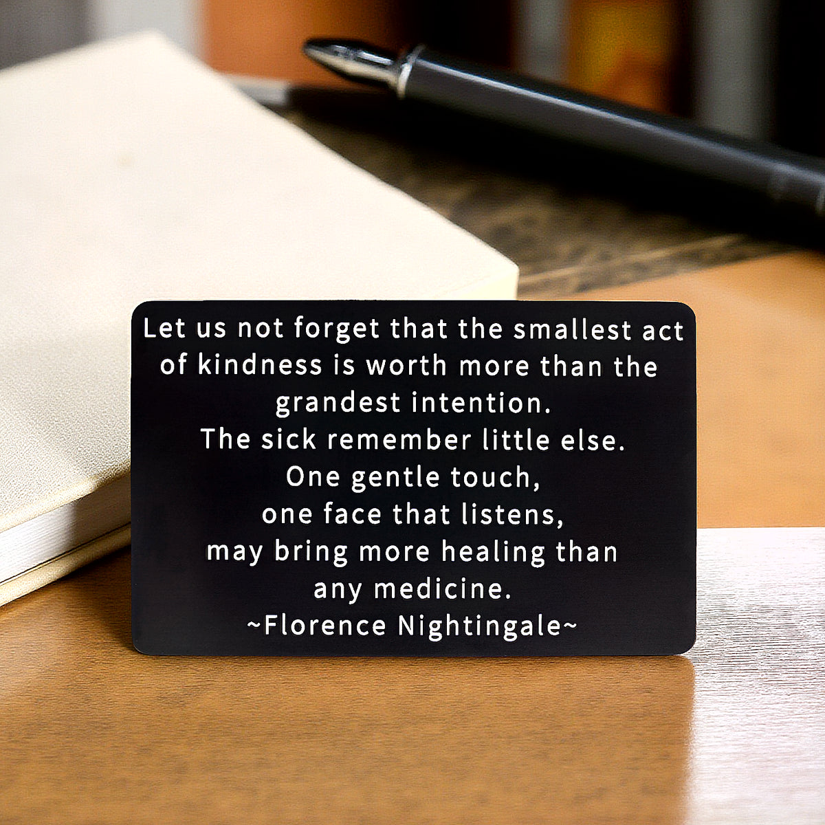Black engraved metal wallet card with Florence Nightingale's quote on kindness and healing, on a table with a white leather-bound journal and black pen.