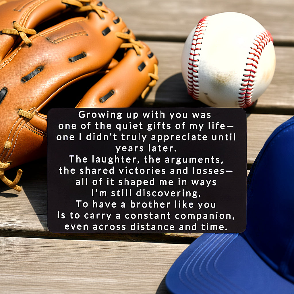 Black engraved metal wallet card reading 'Growing Up With You Was One Of The Quiet Gifts Of My Life' lying on a picnic table next to a baseball, baseball glove, and blue hat.