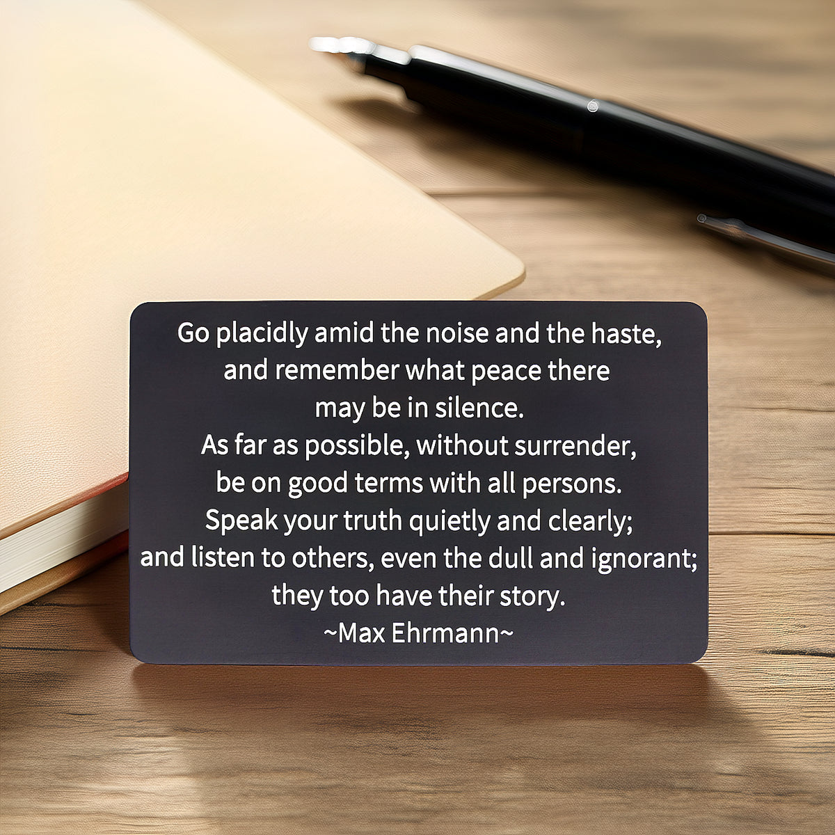 Black engraved metal wallet card with an excerpt from Max Ehrmann's "Desiderata," reading 'Go placidly amid the noise and the haste, and remember what peace there may be in silence' lying on a worn wooden table with a tan leather-bound journal and black pen in the background.