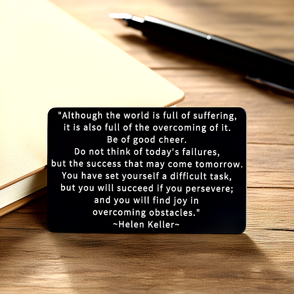 Black engraved metal wallet card with Helen Keller's quote on overcoming suffering, on a worn wooden table with a tan leather-bound journal and black pen.