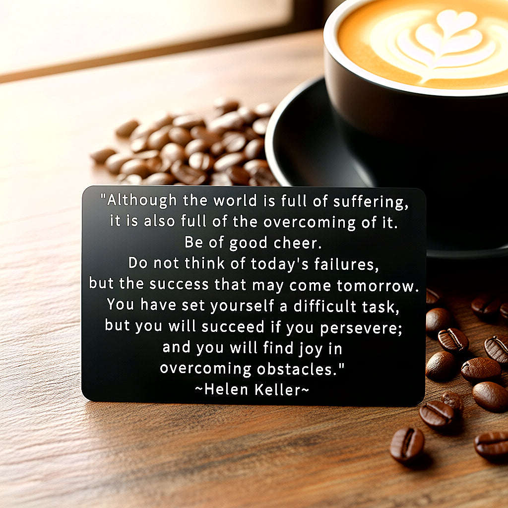 Black engraved metal wallet card with Helen Keller's quote on overcoming suffering, on a wooden table with scattered coffee beans and a cup of coffee.