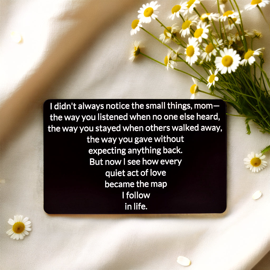 Black engraved metal wallet card reading 'Quiet Acts of Love Map My Life' lying on white linen next to daisies.