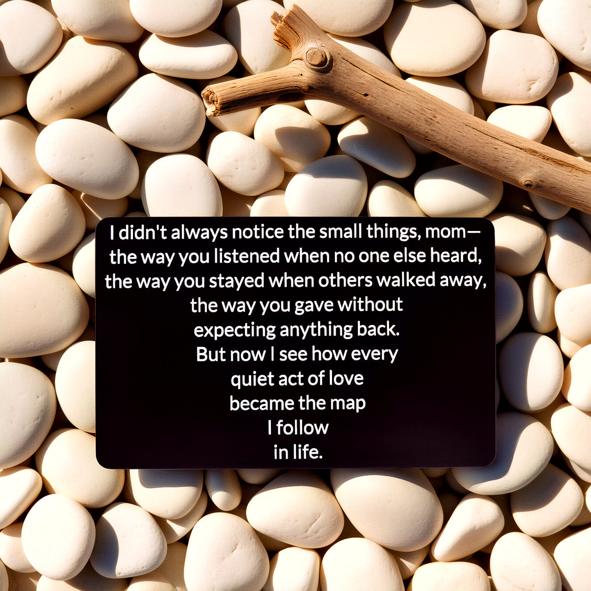 Black engraved metal wallet card reading 'Quiet Acts of Love Map My Life' lying on white river rocks next to a brown stick.