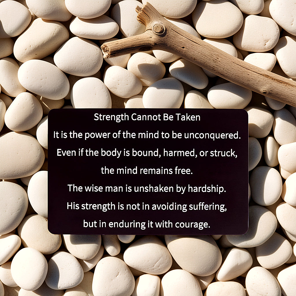 Black engraved metal wallet card reading 'Strength Cannot Be Taken' lying on smooth white river rocks next to a wooden stick.