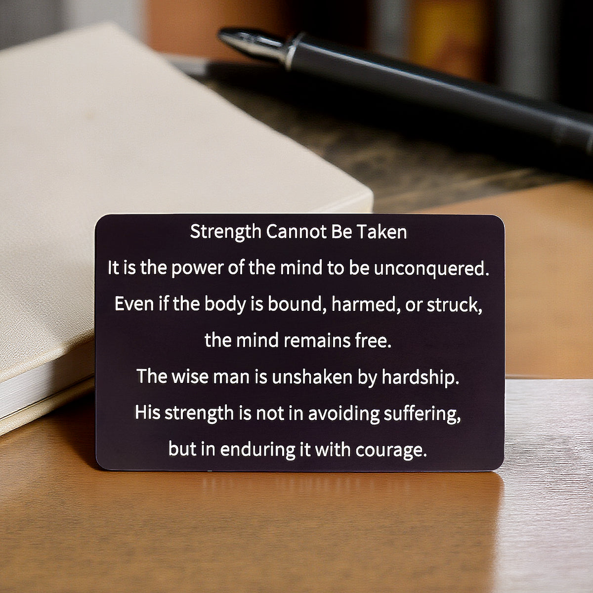 Black engraved metal wallet card reading 'Strength Cannot Be Taken' lying on a wooden table with a white notebook and black pen in the background.