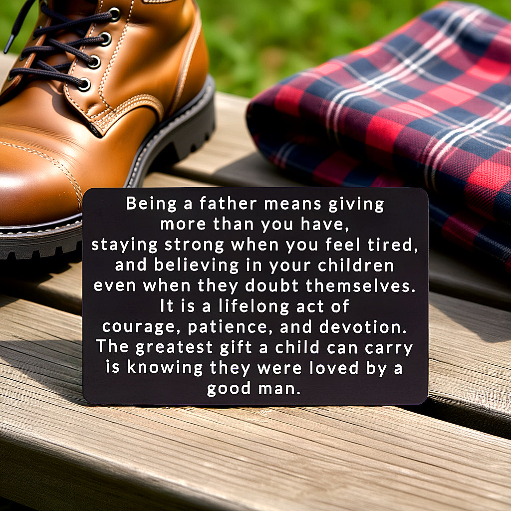 Black engraved metal wallet card with a message about being loved by a good father, lying on a wooden bench next to a brown leather boot and a plaid flannel shirt.