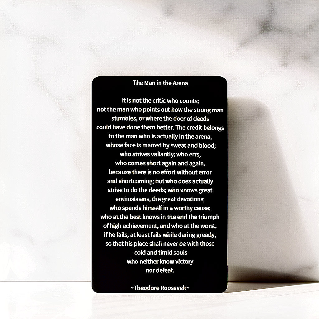 Black engraved metal wallet card reading 'The Man In The Arena,' by Theodore Roosevelt lying on a white grey marbled background.