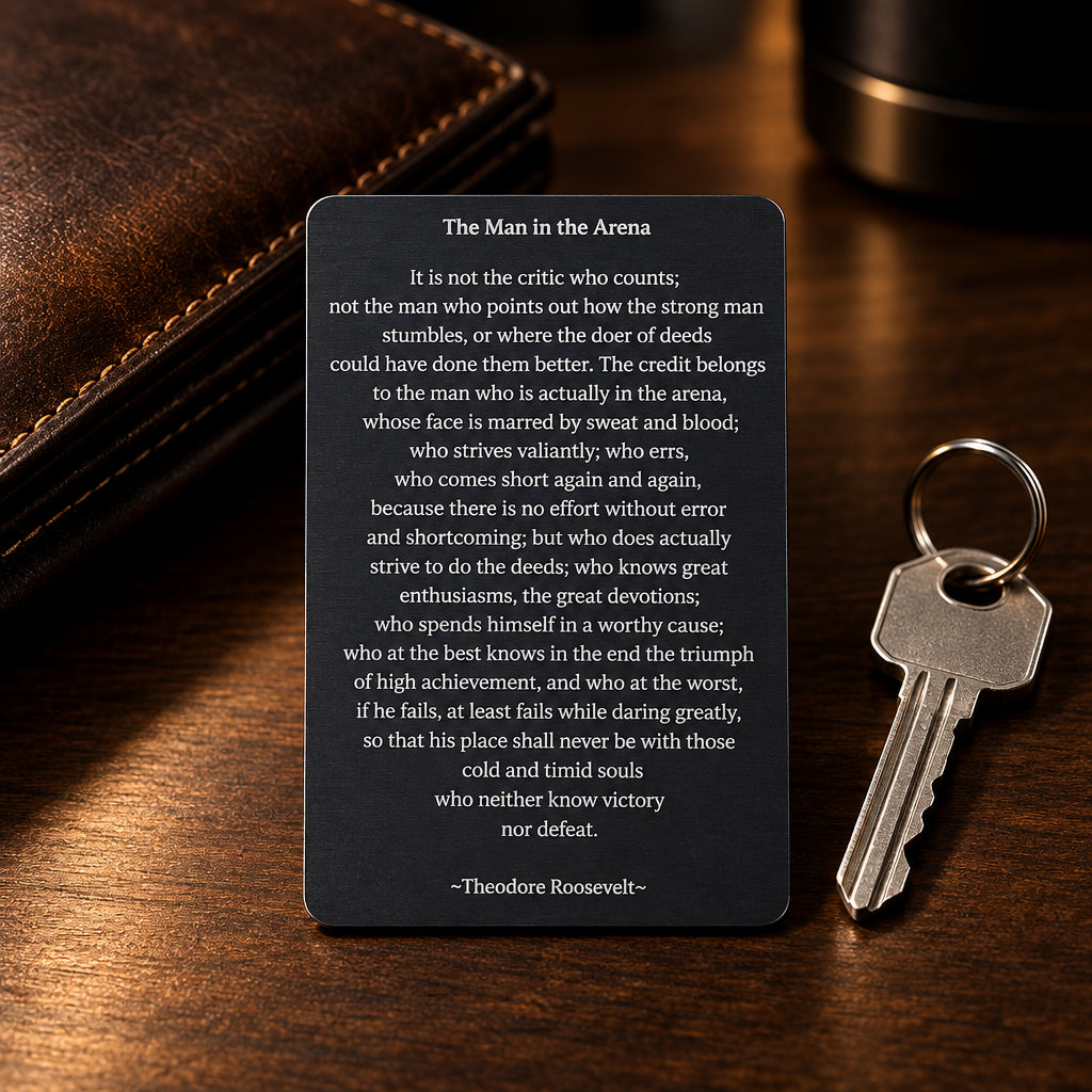 Black engraved metal wallet card reading 'The Man In The Arena,' by Theodore Roosevelt lying on a wooden table next to a brown leather wallet and silver key.