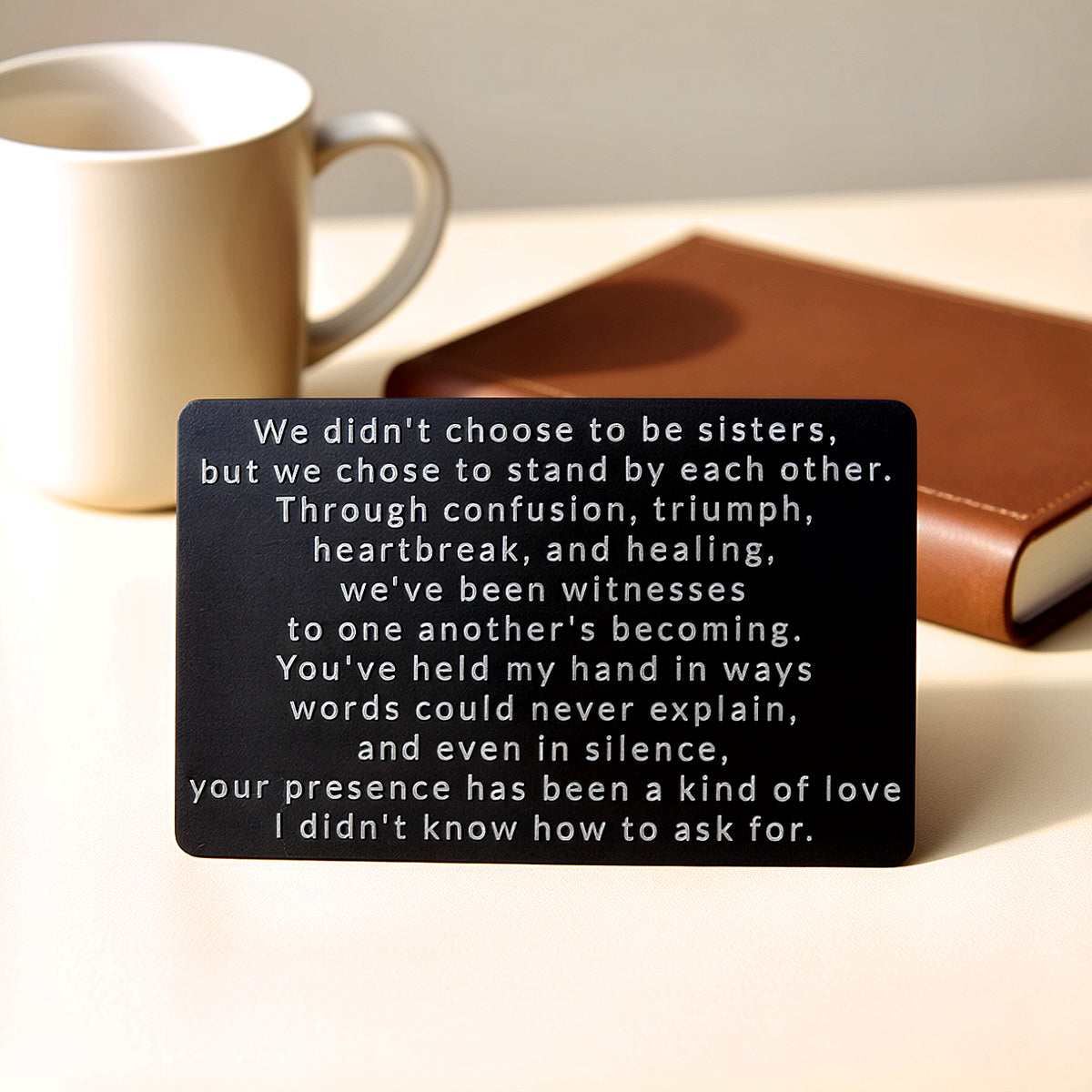 Black engraved metal wallet card with a message about sisters who chose to stand by each other, lying on a table next to a white mug and a brown leather journal.
