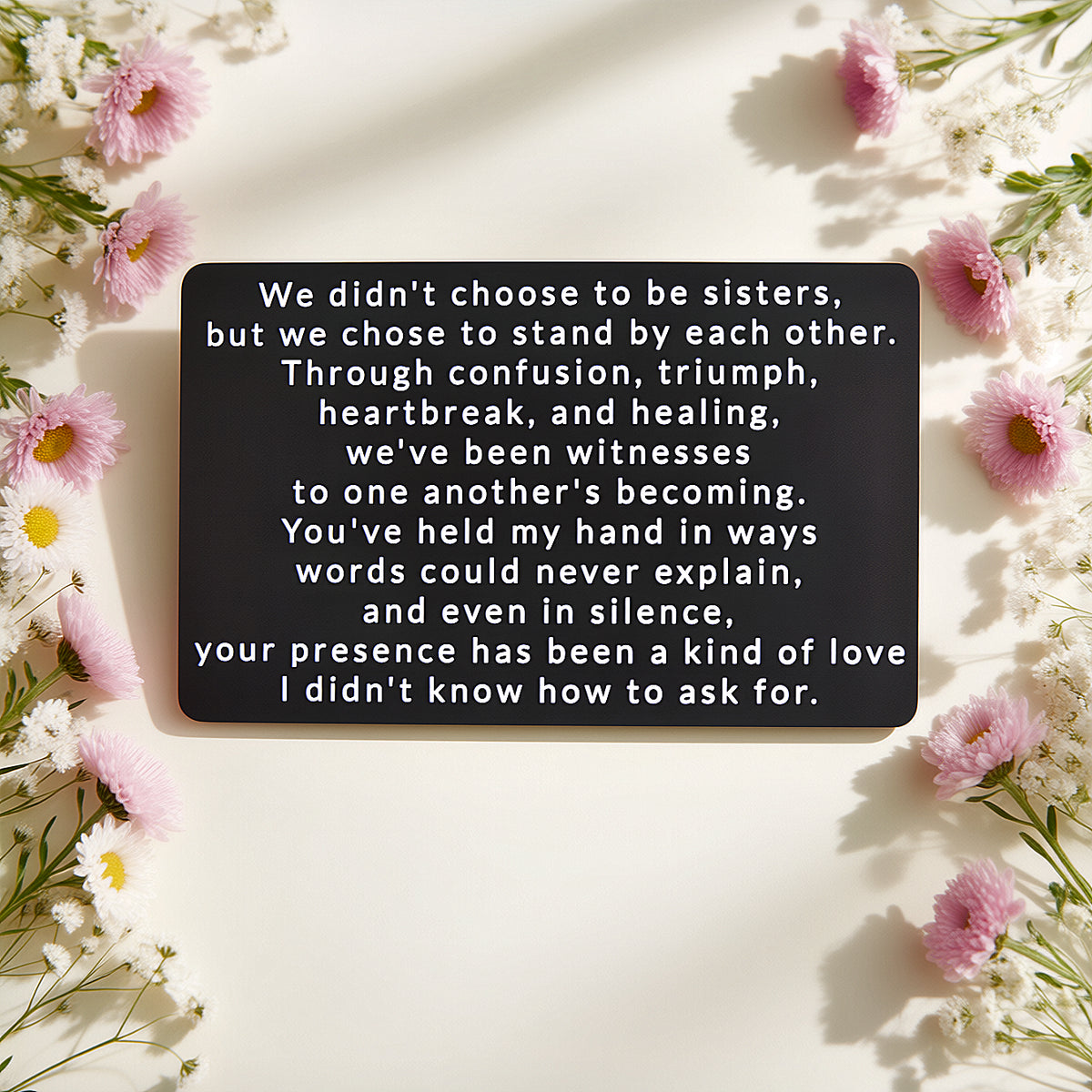 Black engraved metal wallet card with a message about sisters who chose to stand by each other, lying on a cream background framed by pink and white wildflowers.