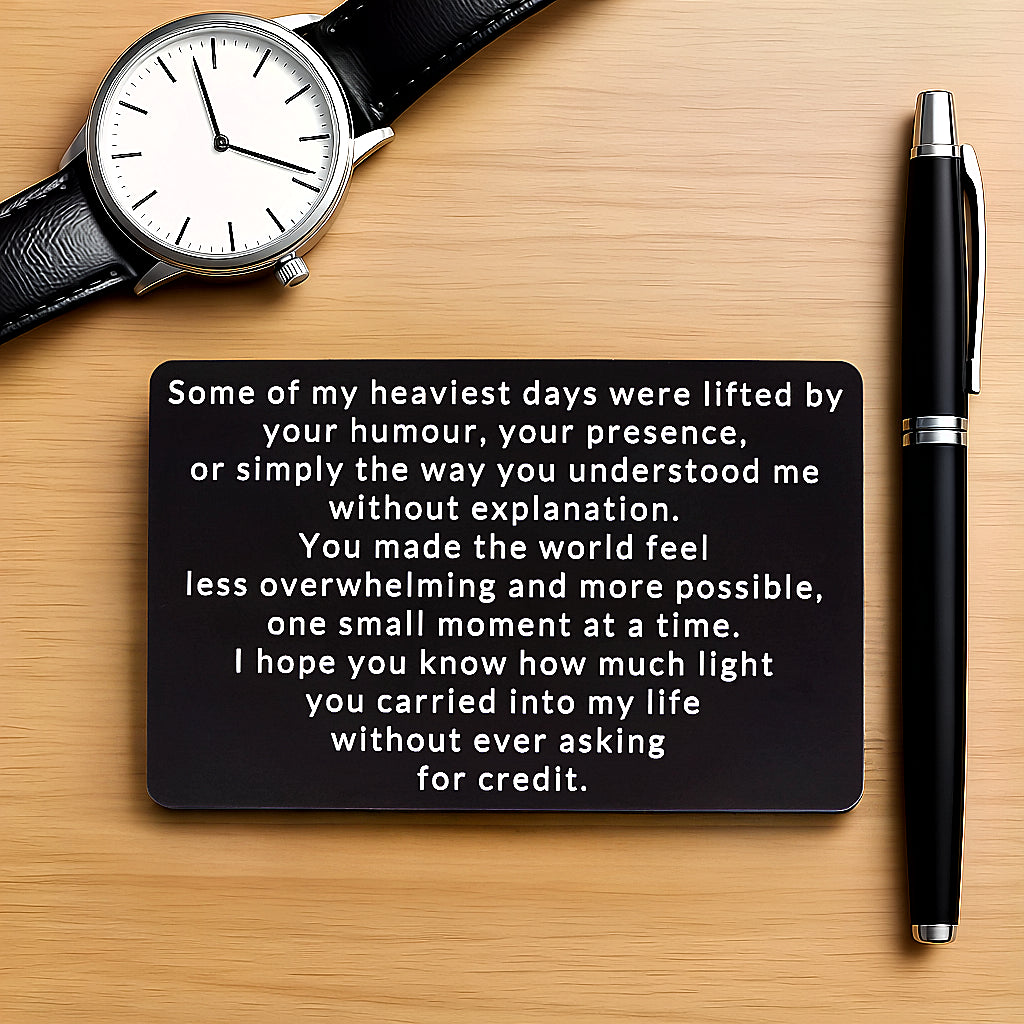 Black engraved metal wallet card reading 'You Brought Light Into My Life Without Asking For Credit' lying on a wood table next to a watch and a pen.