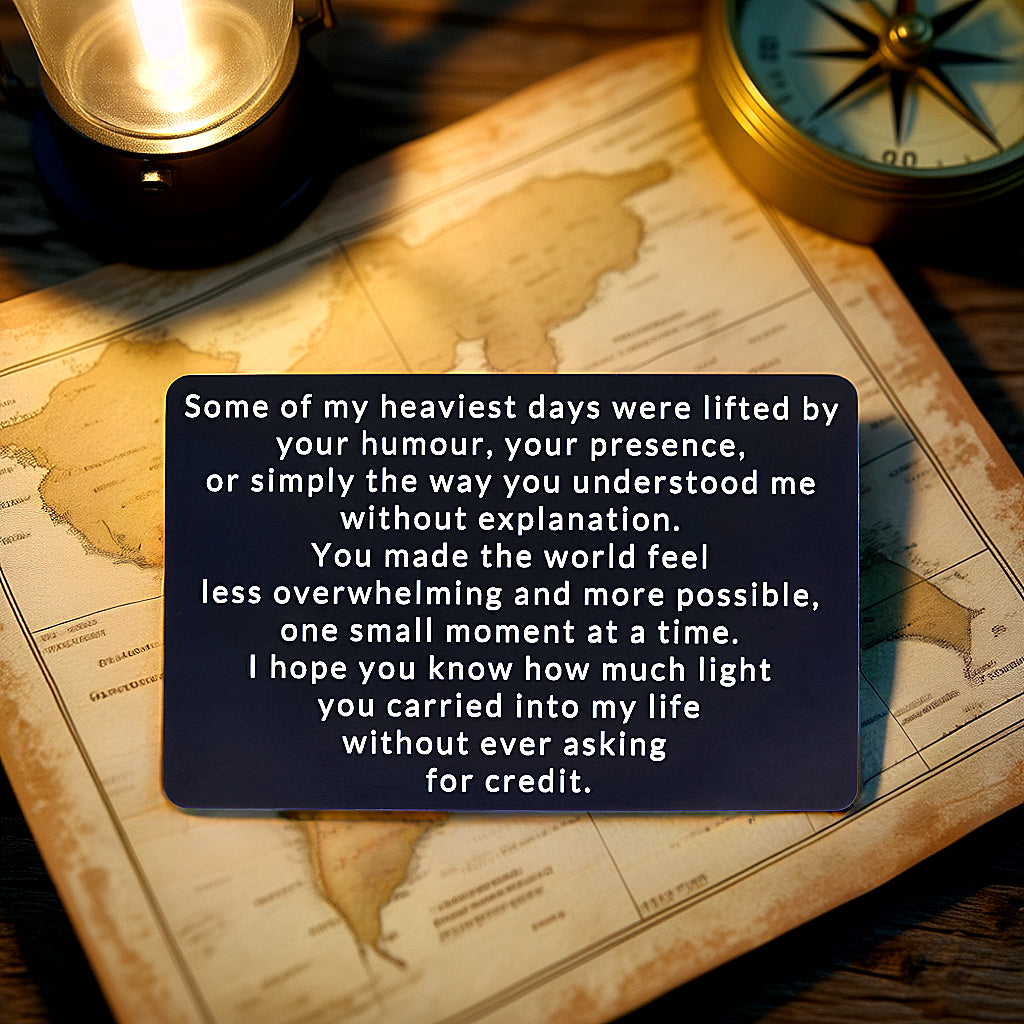 Black engraved metal wallet card reading 'You Brought Light Into My Life Without Asking For Credit' lying on a map next to a compass and lantern.