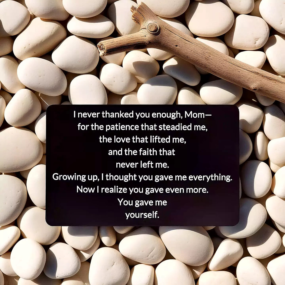 Black engraved metal wallet card reading 'You Gave Me Yourself' lying on a bed of white river rocks next to a wooden stick.