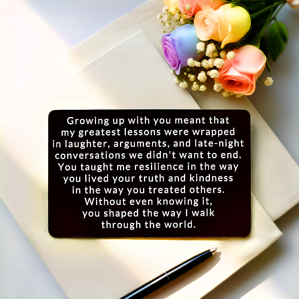 Black engraved metal wallet card reading 'You Shaped the Way I Walk Through the World' lying on a white canvas next to a bouquet of roses and a pen.