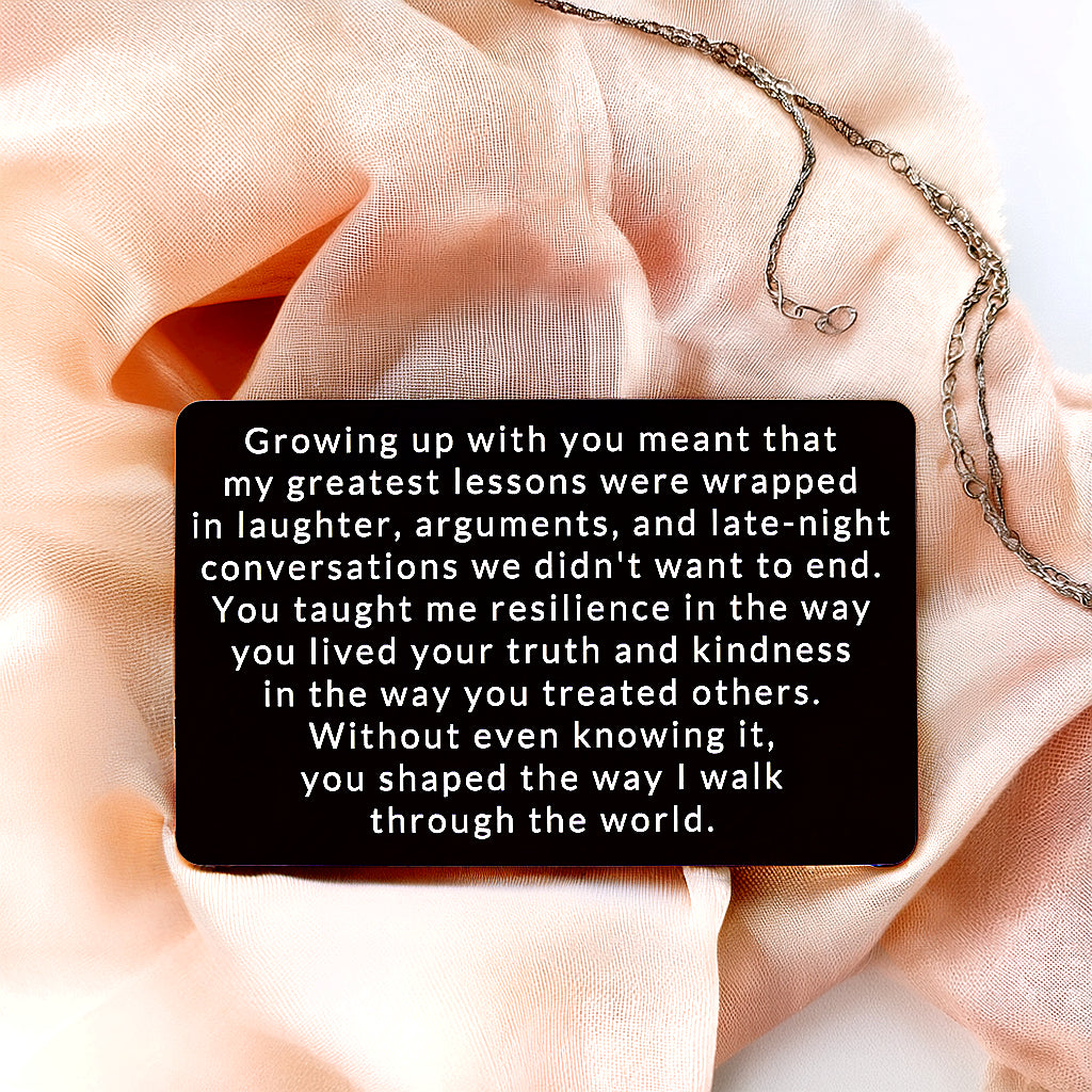 Black engraved metal wallet card reading 'You Shaped the Way I Walk Through the World' lying on blush silk fabric next to a delicate chain necklace.